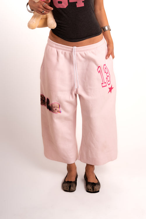 A person wearing light pink cropped sweatpants with a pink '13' graphic and a small dog patch on the left leg, paired with a black graphic top and black flats, against a white studio background.
polar fleece sweatpants | LA Apparel sweatpants | pink graphic sweatpants | puppy print sweats | varsity number sweatpants | oversized fleece sweatpants | cozy loungewear | Y2K sweatpants | dog lover sweatpants | wide leg sweatpants